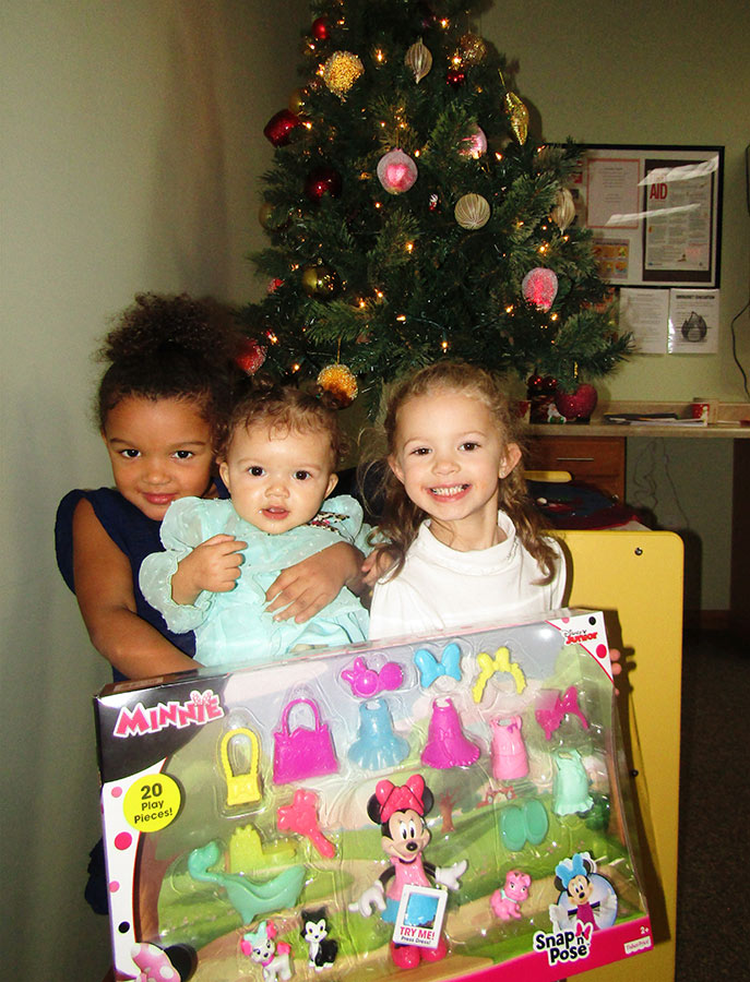 three young girls posing with gift in front of christmas tree